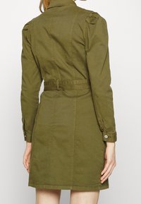 Abito verde oliva realizzato in tessuto leggero, con silhouette aderente, maniche lunghe con polsini abbottonati e vita con cintura.