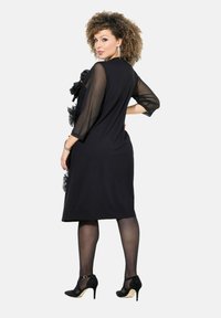 Frau mit lockigem Haar, die ein schwarzes knielanges Kleid mit durchsichtigen Ärmeln und schwarzen High Heels trägt, posiert mit der Hand auf der Hüfte.