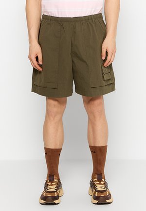 Homme portant un short cargo vert olive avec des poches, des chaussettes Nike marron et des baskets multicolores marron et beige, debout devant un fond blanc.