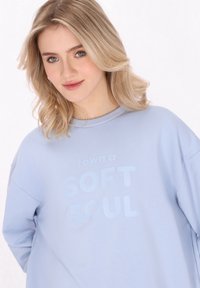 Felpa oversize blu chiaro con una consistenza morbida che presenta il testo "Possiedo un'ANIMA MORBIDA" in un blu leggermente più scuro. Collo rotondo e spalle cadenti.