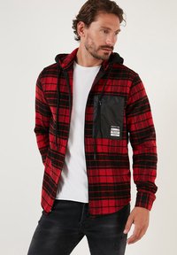 Punainen ja musta ruudullinen flanellitakki hupulla, jossa on musta vetoketjullinen rintatasku ja säädettävät nyörit.
