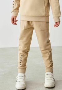 Beige sweatshirt og matchende joggers med teksturerede detaljer og sidelommer. "BAKER" trykt i sort på joggersne. Hvide sneakers.