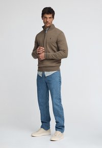 Brun zip-up tröja med muddade ärmar, lager-på-lager över en ljusblå skjorta, kombinerad med ljusblå jeans och cremefärgade sneakers. Enfärgad bakgrund.