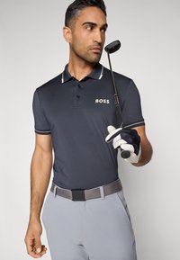 Σκούρο μπλε ναυτικό πόλο με λευκές λεπτομέρειες, με λογότυπο στο στήθος. Γκρι ζώνη και ανοιχτό γκρι παντελόνι, κρατώντας ένα γκολφ κλομπ.