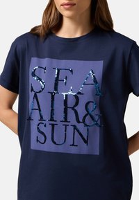 T-shirt en coton bleu marine présentant un design pailleté avec le texte "AIR DE MER & SOLEIL" dans des textures et des nuances variées de bleu et de noir.