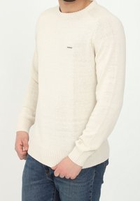 Cremfarbener Strickpullover mit gerippten Bündchen und Saum, versehen mit einem kleinen Markenlogo auf der Brust. Die Stofftextur wirkt weich und leicht.