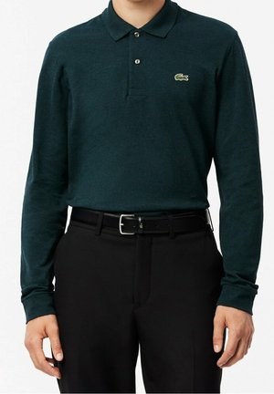 Homme portant un polo vert foncé à manches longues avec logo crocodile, ceinture noire et pantalon noir, debout sur fond blanc.