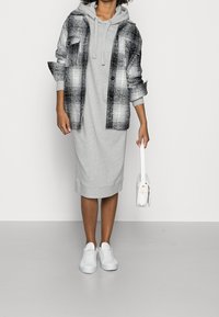 Mujer vistiendo un vestido gris con capucha, chaqueta de cuadro blanco y negro, zapatillas blancas, sosteniendo un pequeño bolso blanco contra un fondo liso.