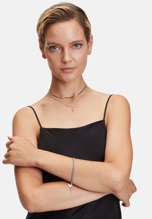 Silberne Schmuckstücke mit geschichteten Ketten, einem geschwungenen Choker und einem Armband mit einem Herzanhänger auf einem glatten schwarzen Kleid.