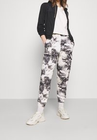 Svart lättviktsjacka med dragkedja, kombinerad med en vit kort t-shirt, tillsammans med mönstrade sweatpants i svart och krämfärgad. Vita sneakers.