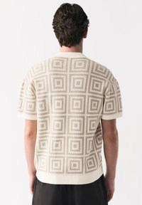 Mann von hinten gesehen, trägt ein beige kurzes Strickoberteil mit einem geometrischen Quadratmuster und schwarze Hose.