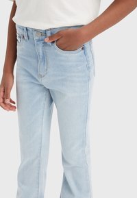 Osoba v světle modrých džínách, s jednou rukou v přední kapse, a s bílou košilí zastrčenou uvnitř.