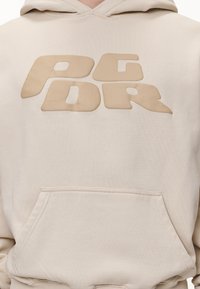 Beige mikina s kapucí, s přední kapsou a zaoblenými, vystouplými písmeny "AGDR" ve velkém, stylizovaném písmu na hrudi.