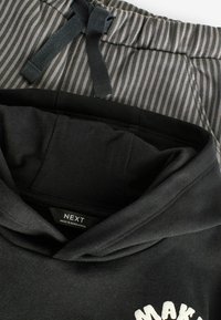 Sort hættetrøje med hvid tekst, parret med gråstribede sweatpants. Hætten og snøre detaljerne er synlige, hvilket fremhæver et afslappet design.
