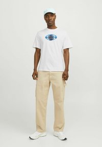 Hvid bomulds T-shirt med blåt grafisk logo, kombineret med beige cargo-bukser og hvide løbesko. Lys blå cap fuldender outfittet.