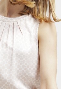 Hihaton vaaleanpunainen paita, jossa on hienovarainen geometrinen kuvio ja pehmeitä laskoksia kaulan läheisyydessä, henkilöllä on vaaleat laineikkaat hiukset.