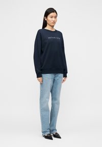 Μπλε σκούρα φούτερ με στρογγυλή λαιμόκοψη και κεντημένο λογότυπο, συνδυασμένο με ανοιχτό μπλε τζιν. Το μοντέλο στέκεται με τα χέρια στα πλευρά, φορώντας μυτερά μαύρα παπούτσια.