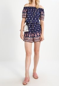 Combinaison off-shoulder bleu marine avec des motifs floraux, encolure élastique et shorts. Associée à des sandales à glisser roses et un petit sac bandoulière en argent.