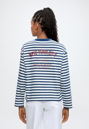 Femme aux cheveux tressés porte une chemise à manches longues rayée bleue et blanche avec le texte "New York City" dans le dos et un pantalon blanc.