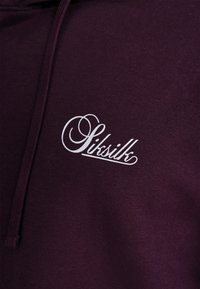 Tmavě fialová mikina s kapucí s bílým nápisem ve smyčkovém designu. Materiál vypadá měkký s texturovaným povrchem a nastavitelnou šňůrkou.