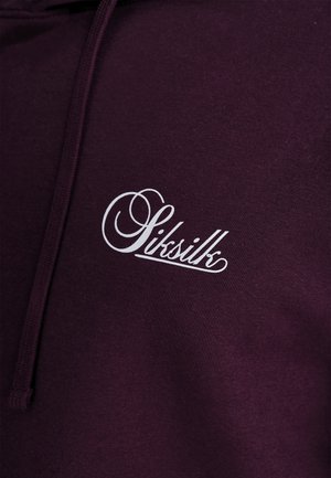 Dunkel-lila Kapuzenpullover mit einem weißen Skriptlogo und einem geschwungenen Design. Das Material wirkt weich mit einer strukturierten Oberflächenbeschaffenheit und hat einen verstellbaren Kordelzug.
