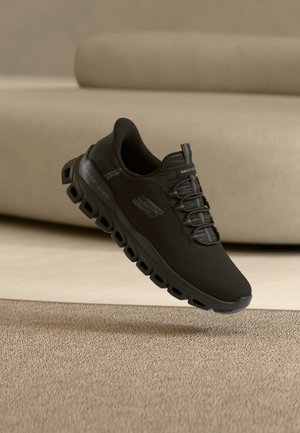 Black Skechers slip-on athletic shoe with breathable mesh upper and cushioned sole suspended above beige carpet and sofa background.
