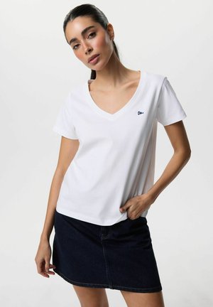 Kobieta o ciemnych włosach, ubrana w białą koszulkę z dekoltem w serek i ciemną dżinsową spódnicę, stojąca z jedną ręką w kieszeni na jednolitym tle.