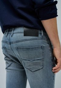 Sive traperice slim kroja, s crnim kožnim označivačem "SALSA JEANS" i istaknutim detaljima šivanja na dzepovima.