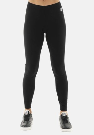 Onderlichaam van een persoon die zwarte leggings en zwarte sneakers met witte zolen draagt, staande tegen een witte achtergrond.