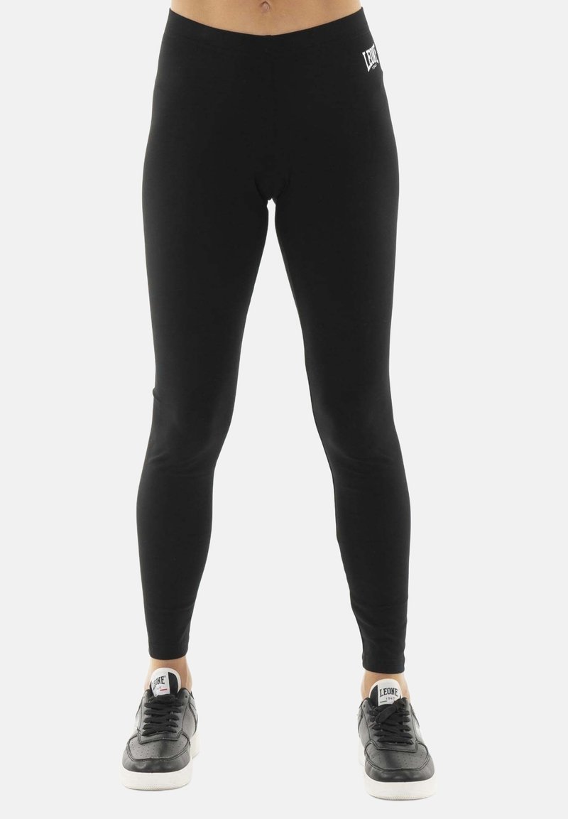 Partie inférieure du corps d'une personne portant des leggings noirs et des baskets noires avec des semelles blanches, debout sur fond blanc.