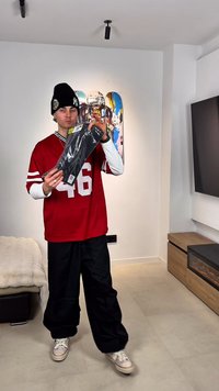 Jeune homme portant un bonnet et un maillot rouge tient un objet noir emballé dans un salon moderne avec une décoration skate sur le mur.