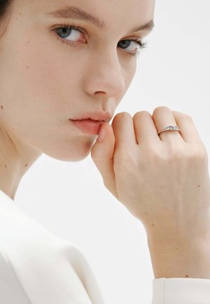Joven mujer con piel clara y ojos azules apoyando la barbilla en el puño, con un anillo de plata con tres piedras preciosas, ropa blanca, fondo neutro.