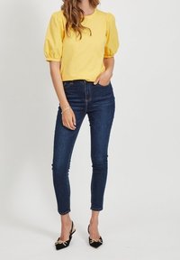 Żółta bluzka z bufiastymi rękawami w połączeniu z dopasowanymi ciemnoniebieskimi dżinsami i czarnymi szpilkami z złotymi akcentami. Białe tło.