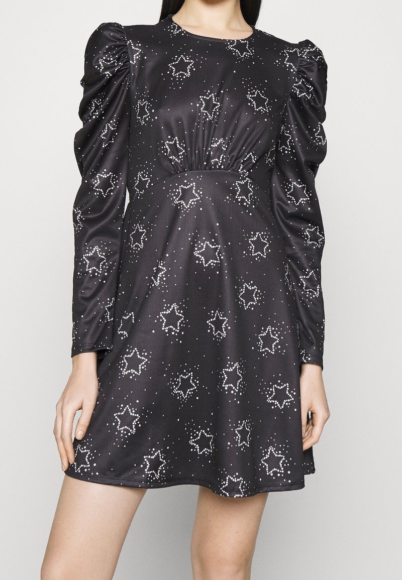 Robe noire à manches longues avec épaules bouffantes et motifs d'étoiles blanches, portée par un mannequin aux bras et jambes visibles.