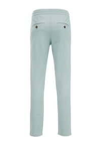 Svetle zelené tepláky vyrobené z měkké látky, s dvěma zadními kapsami s kulatými hnědými knoflíky a elastickým pasem.