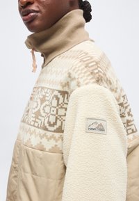 Personne portant un pull en polaire beige et crème à motif avec un col montant et un patch sur la manche indiquant "Kari Traa."