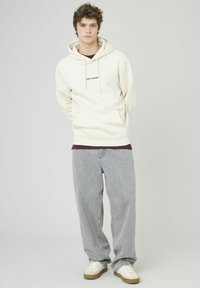 Sweat à capuche crème avec cordons de serrage et poche avant, associé à un jean gris ample et des baskets blanches avec des semelles marron. Design simple, texture douce.