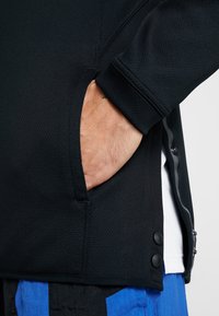 Chaqueta negra texturizada con un bolsillo lateral y botones a presión en el dobladillo. La tela tiene un patrón de malla, que muestra transpirabilidad y flexibilidad.