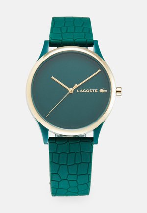 Green rubber watch with a textured strap, gold accents, and a round face featuring a dark green dial with gold hands and logo.