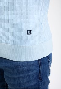 Gaiši zils adīts džemperis ar horizontālu riboto tekstūru un tumši zilu logo apakšējā pusē. Pabeigts ar ribotu apakšmalu.