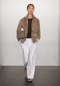 Suède bruine jas met zwarte knopen, bruine top, witte wijde broek en zwarte slippers. Model staat in een minimalistische omgeving.