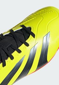 Zapatilla deportiva de color amarillo brillante con superficie texturizada, cordones negros y tres rayas negras. Presenta detalles suaves y estampados.