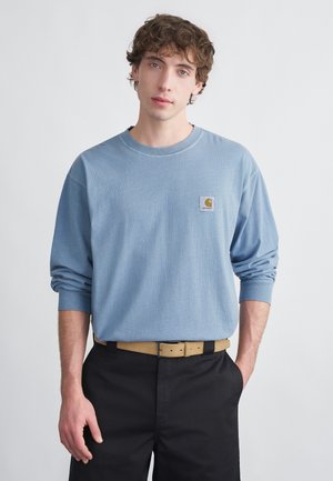 Gaiši zila garām piedurknēm krekls ar ribotiem aproču galiem un nelielu taisnstūrveida logotipa plāksteri; kombinēts ar melnām biksēm un gaiši brūnu jostu.