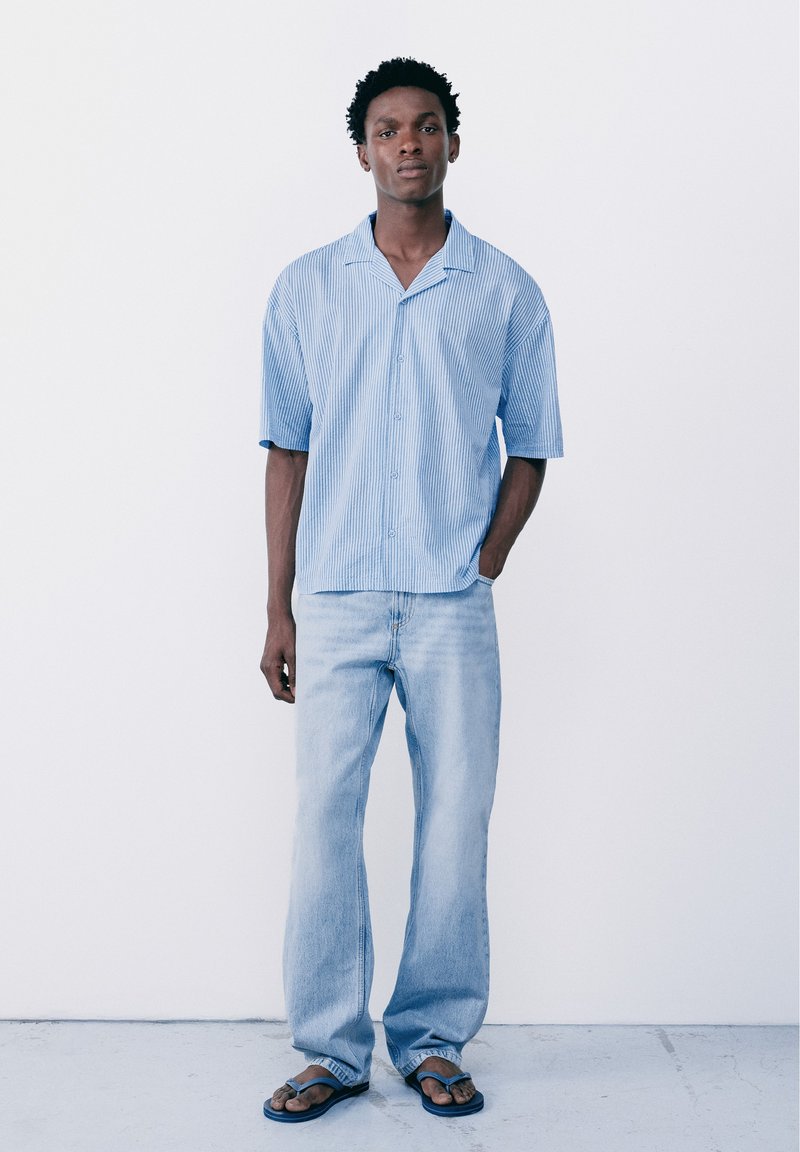 Jeune homme debout, une main dans la poche, portant une chemise à manches courtes rayée bleu clair, un jean clair délavé et des tongs foncées sur fond blanc.