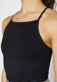 Femme portant un haut noir sans manches, ajusté, avec des fines bretelles et un motif côtelé texturé autour de la taille.