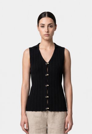 Donna con capelli scuri raccolti indossa un top nero senza maniche a coste con bottoni curvi unici e pantaloni beige chiari, in piedi davanti a uno sfondo semplice.