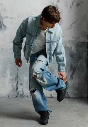 Jeune homme en veste en jean claire et jeans déchirés ajustant sa basket noire devant un mur gris texturé.