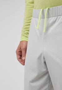 Hellgraue Sporthose aus glattem, leichtem Material mit einem neongrünen Kordelzugbund. Nahaufnahme der Hand auf dem Stoff.
