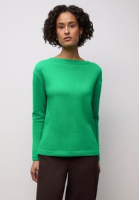 Pull vert en maille avec un large col, des manches longues et des poignets côtelés. Porté avec un pantalon marron, affichant une texture lisse.