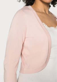 Light pink cardigan with a smooth texture, cropped design, open front, and long sleeves. Features subtle stitching along the edges.